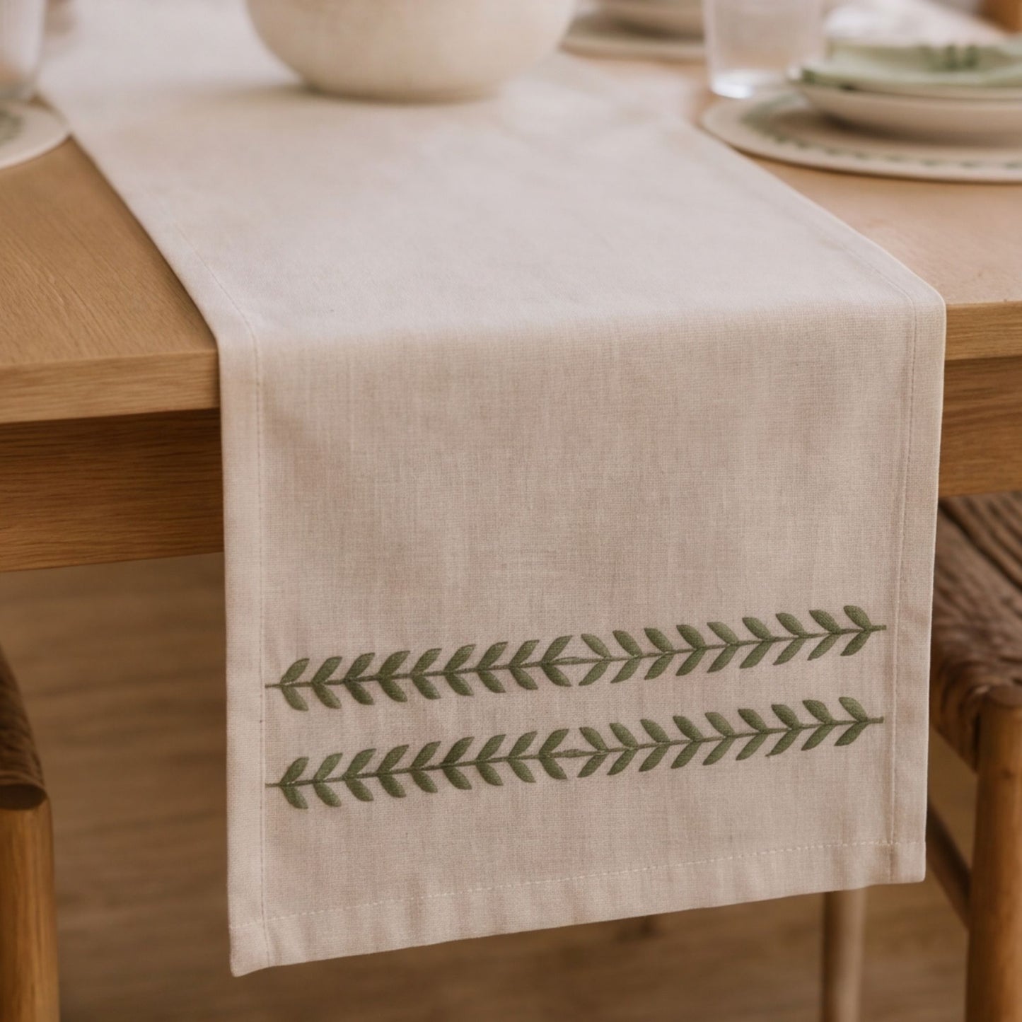 A light beige linen table runner with two horizontal rows of green embroidered leaf patterns, draped over a wooden dining table.