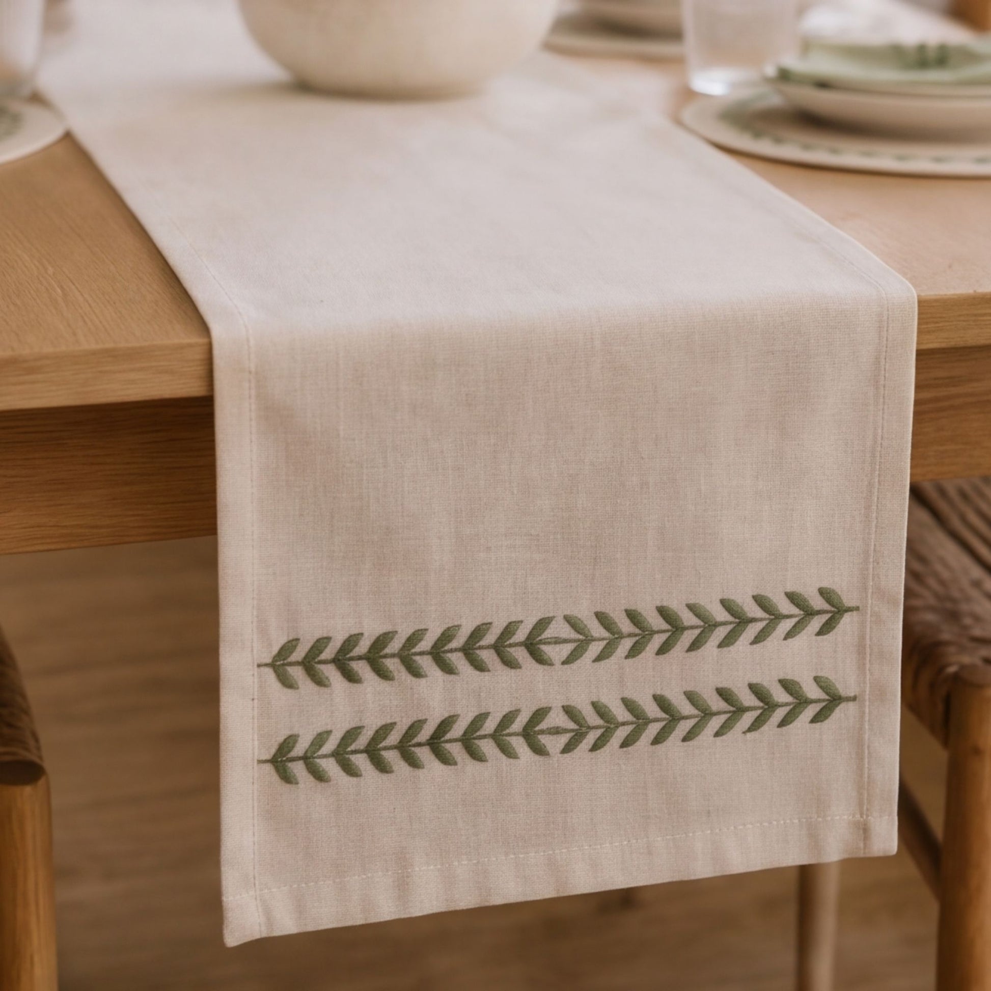 A light beige linen table runner with two horizontal rows of green embroidered leaf patterns, draped over a wooden dining table.
