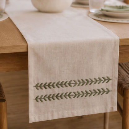 A light beige linen table runner with two horizontal rows of green embroidered leaf patterns, draped over a wooden dining table.