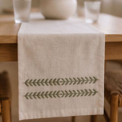 A beige linen table runner with two horizontal rows of green embroidered leaf patterns, draped over a wooden dining table.