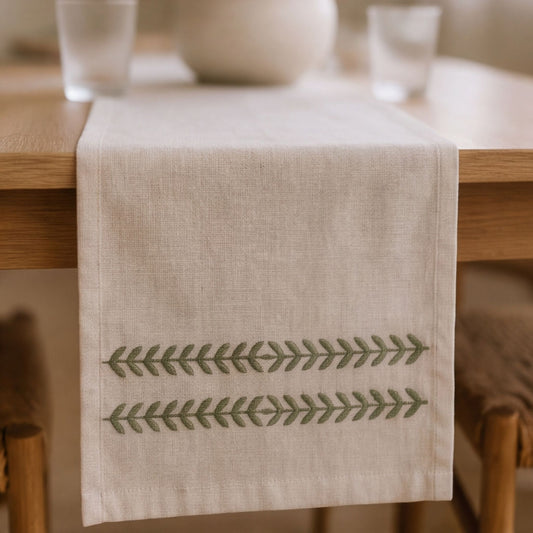 A beige linen table runner with two horizontal rows of green embroidered leaf patterns, draped over a wooden dining table.